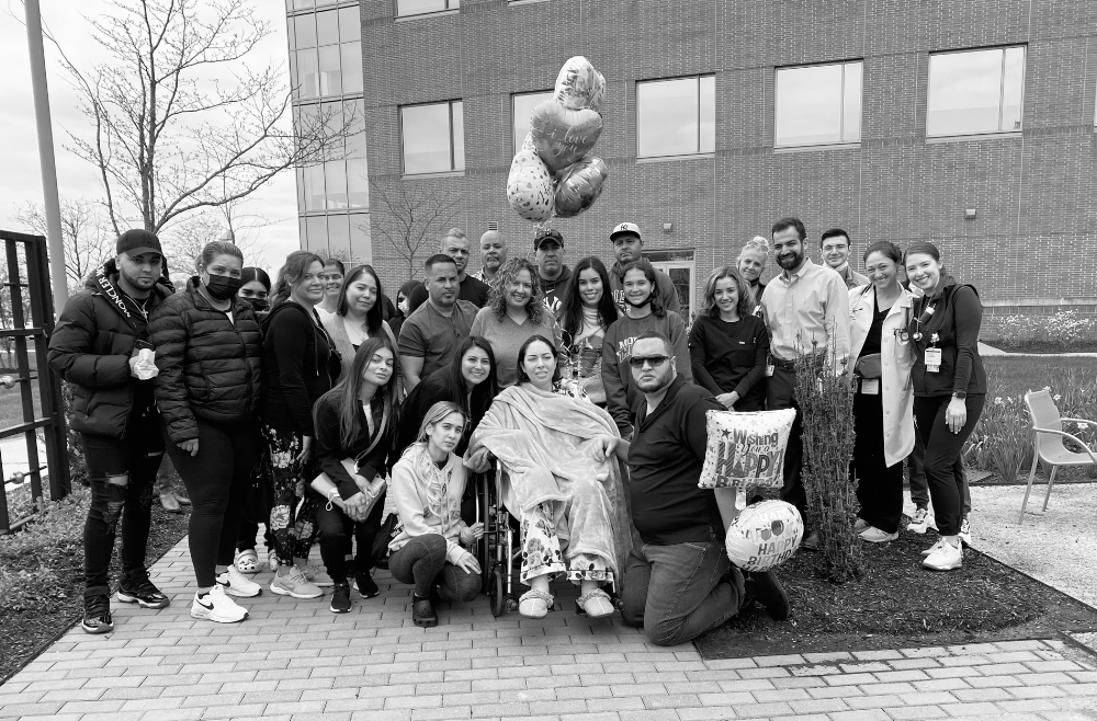 A patient sits in a wheelchair outside, surrounded by a crowd of people, some of whom hold balloons.